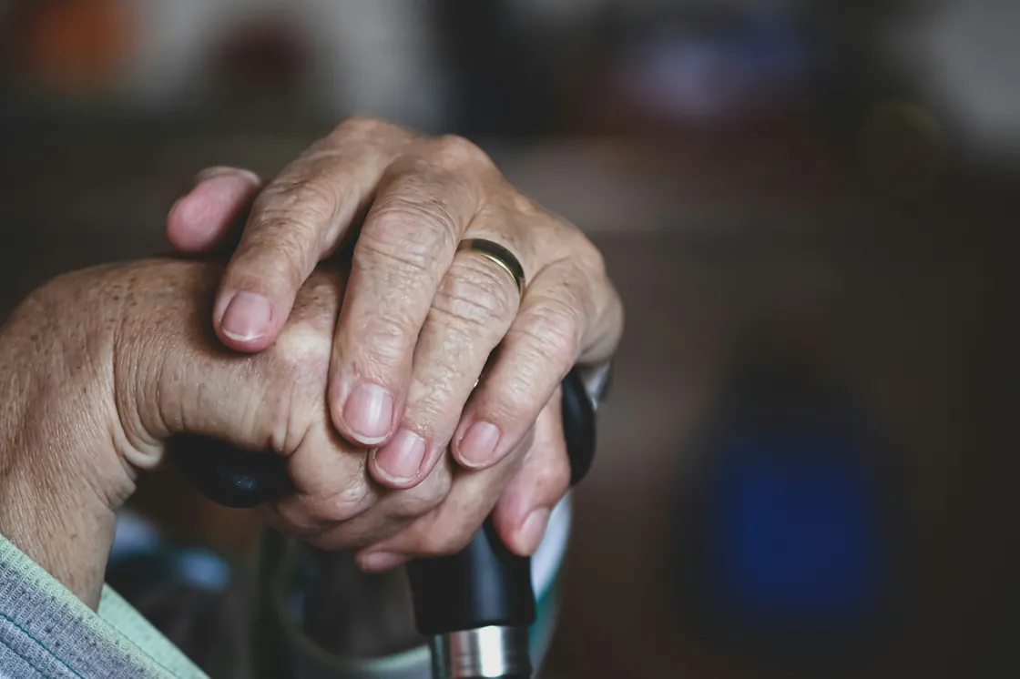 An elderly person's hands resting on a walking stick, with a carer's hand gently supporting them